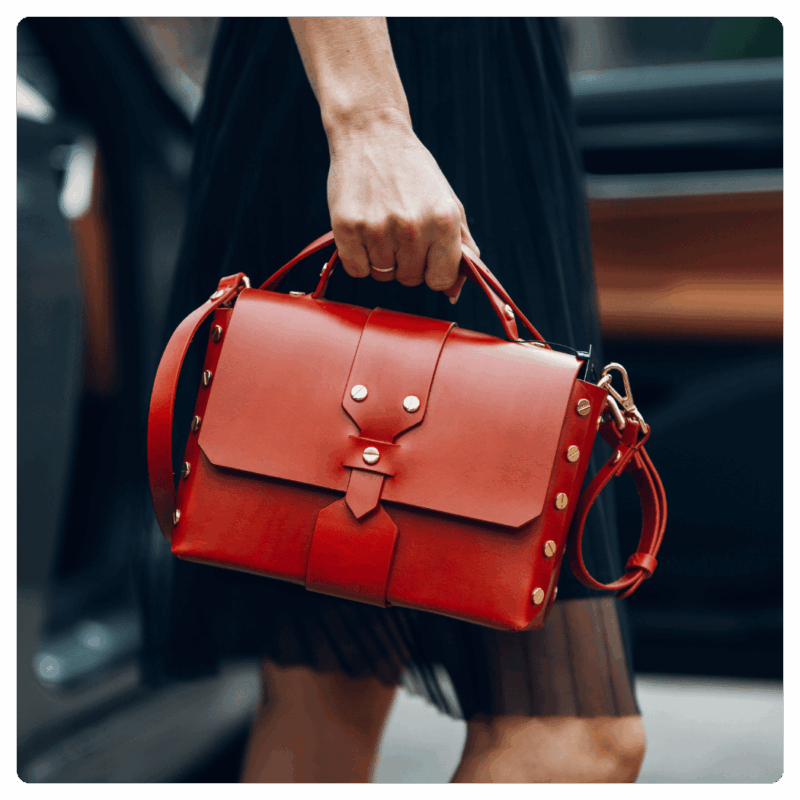 Luxury red leather handbag with gold rivets, top handle, and detachable strap, carried by a woman in a black pleated skirt — elegant fashion accessory for modern style.