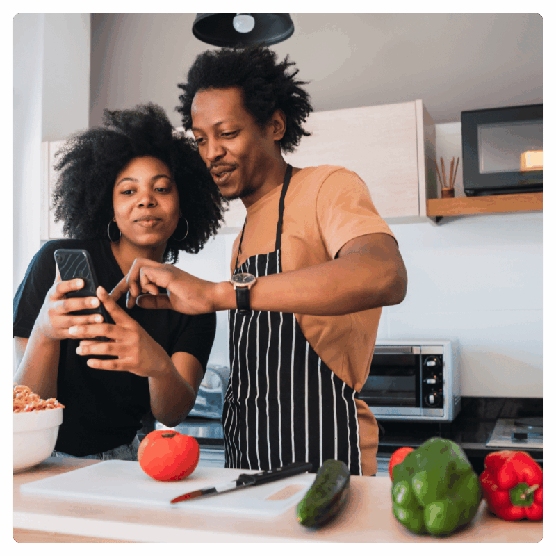 A couple in a kitchen looks at a smartphone while preparing fresh vegetables together.
